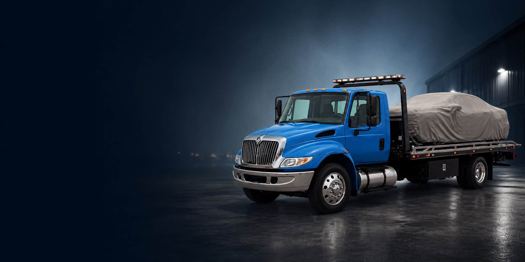 Blue rollback tow truck in an industrial yard at dusk