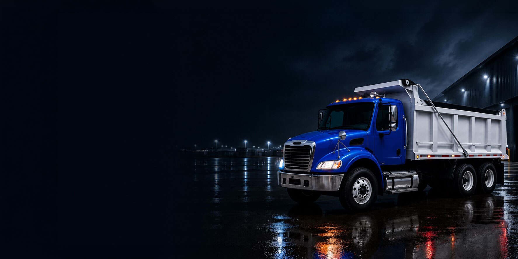 Blue dump truck at night beside an industrial building on wet pavement