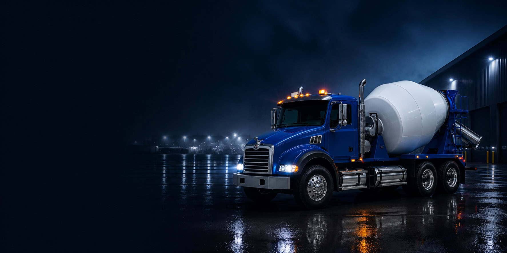 Blue ready-mix concrete truck at night beside an industrial building on wet pavement