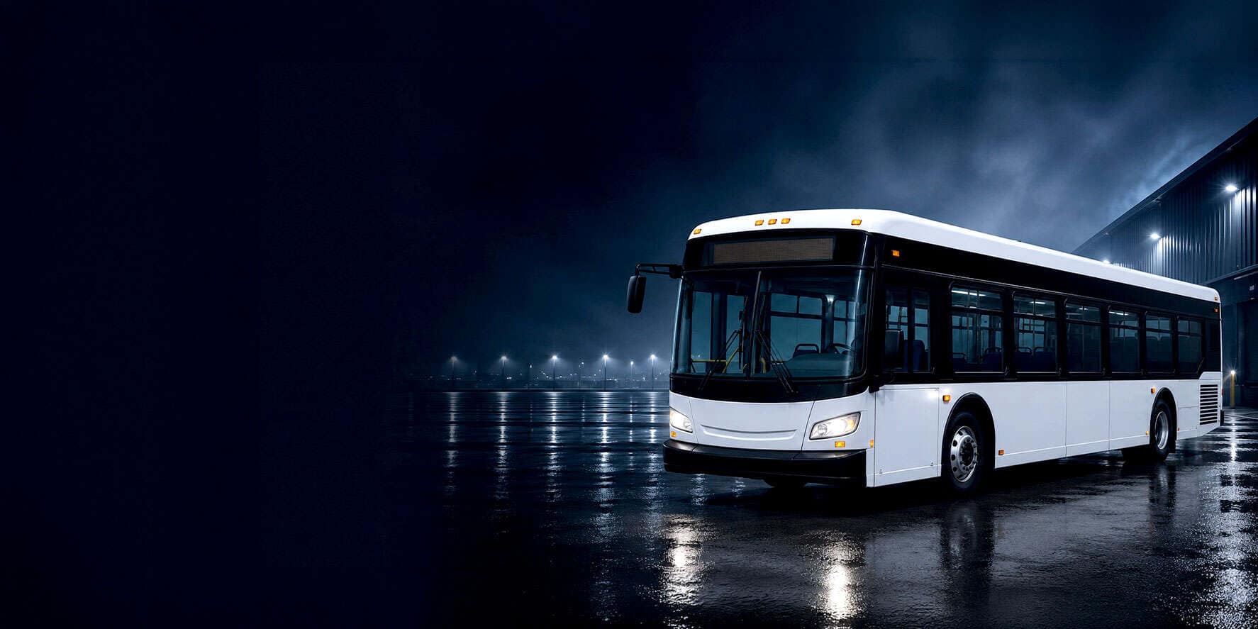 White transit bus at night beside an industrial building on wet pavement